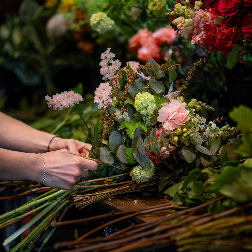 Création Florale : Bouquet Frais Rose et Vert Artisanale Mains d'un floriste composant un bouquet frais : œillets roses, petites fleurs pâles, eucalyptus et hydrangéas verts, sur un établi en bois.
